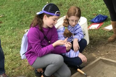 A photo of two younger people sifting through some dirt and discovering hidden objects.
