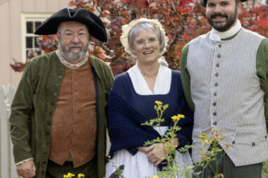 A photo showing three people in period clothing standing next to a sundial and flowers.