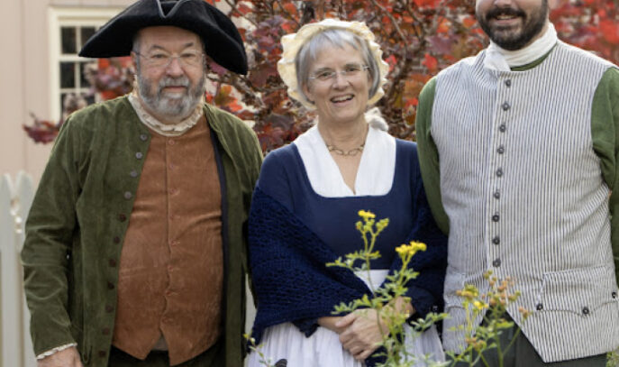 A photo showing three people in period clothing standing next to a sundial and flowers.