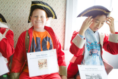 Two children are pictured inside wearing placards around their necks with the information of a historical figure on them. Both are wearing tri-corner hats and red coats of the British Army.