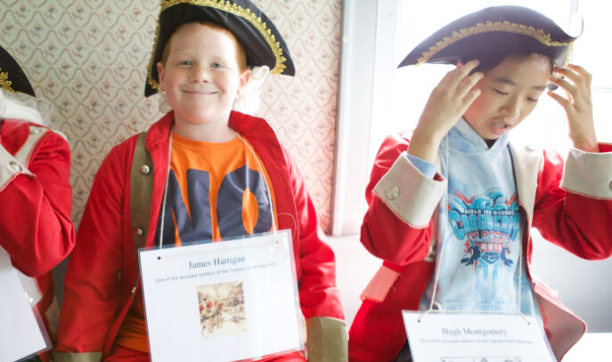 Two children are pictured inside wearing placards around their necks with the information of a historical figure on them. Both are wearing tri-corner hats and red coats of the British Army.