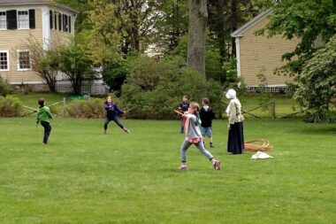 Children playing on a green lawn with a woman in period garb.