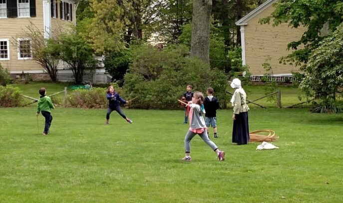Children playing on a green lawn with a woman in period garb.