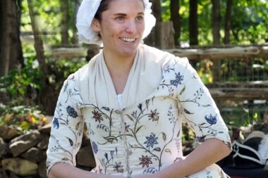 A photo of a woman representing Deborah Sampson. She is wearing a white dress with flowers on it and is smiling. Her hands are clasped in her lap.