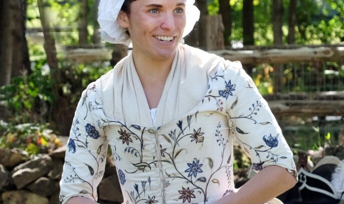 A photo of a woman representing Deborah Sampson. She is wearing a white dress with flowers on it and is smiling. Her hands are clasped in her lap.