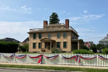 An exterior photo of Quincy House. The white picket fence out front is visible with red, white, and blue bunting.